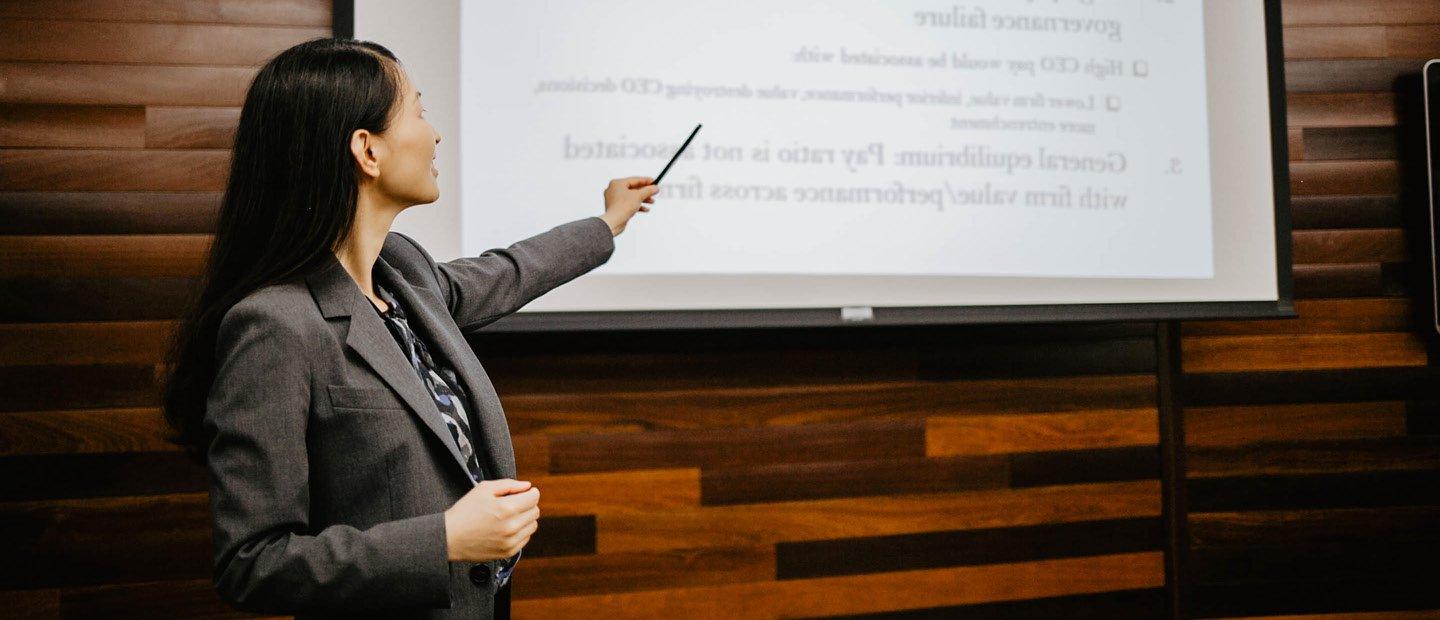 A woman in a suit standing in front of a projector screen, pointing at a line of text on it.