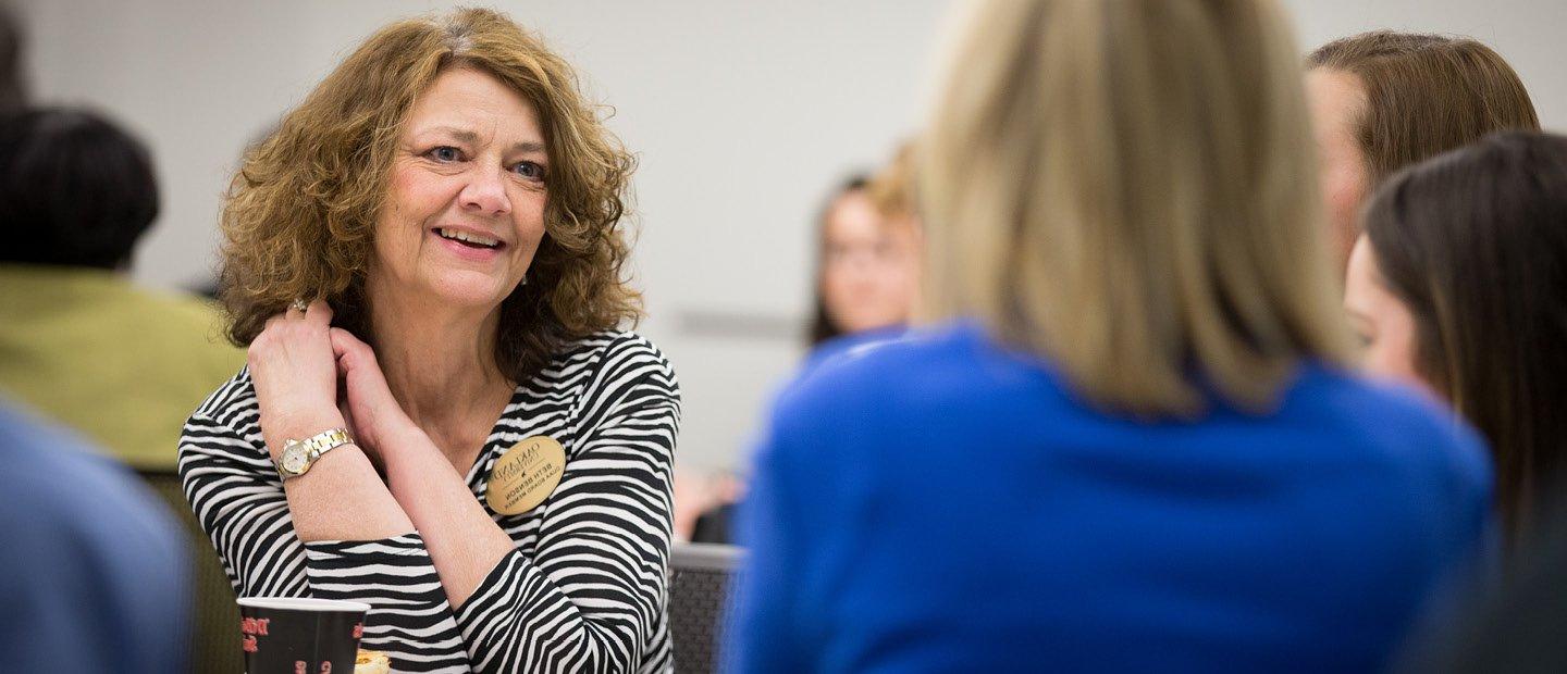 A woman wearing a name tag, seated, talking to three other women.