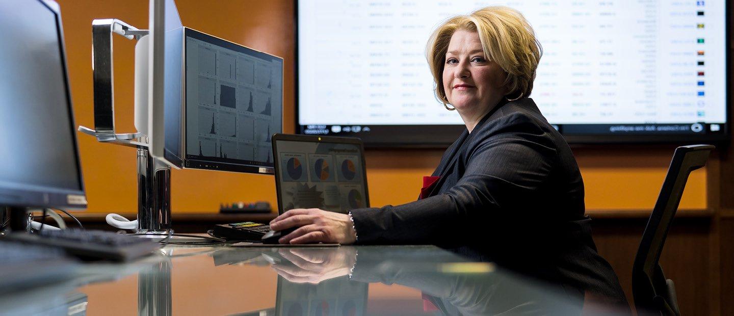 A woman seated a desk with multiple computer screens and a projector behind her, looking at the camera.